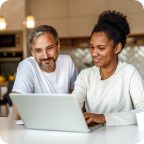 A couple sit in their bright kitchen and use a laptop to shop online together.  